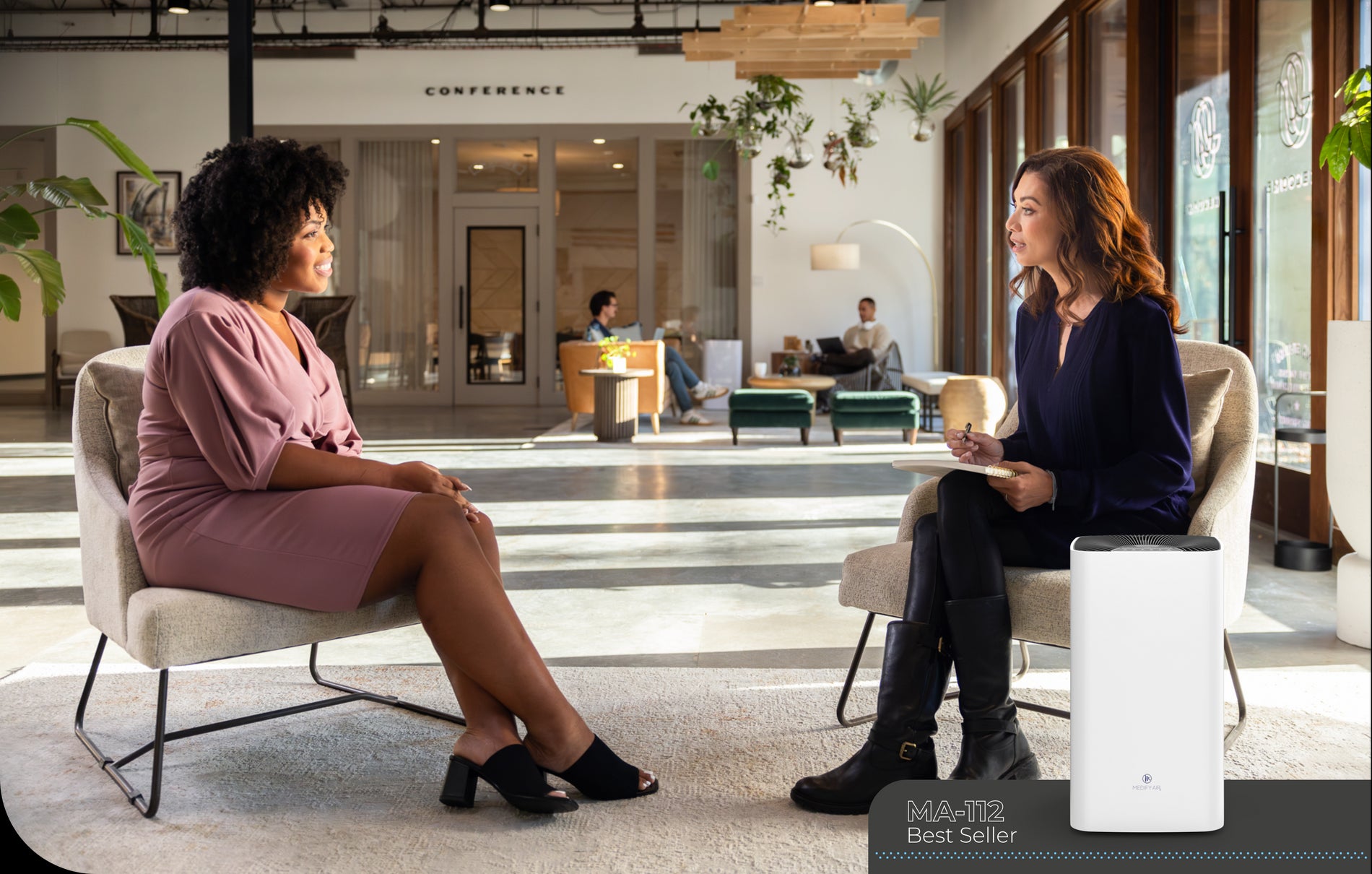 two ladies chatting in an office setting that is brightly lit from natural sun and a MA-112 air purifier