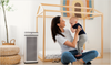 A woman sitting on the floor holds a baby while next to a white and black air purifier, with a wooden house-shaped play gym behind them in a bright room.