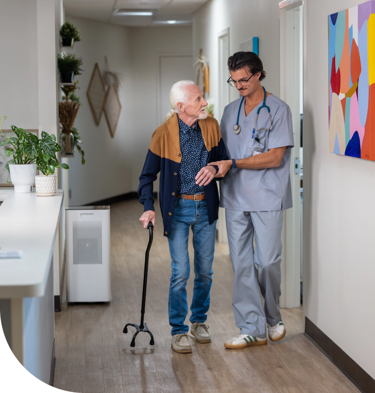a senior citizen and a nurse practitioner walking a hallway of a care facility next to an administration desk that has two plants and an air purifier