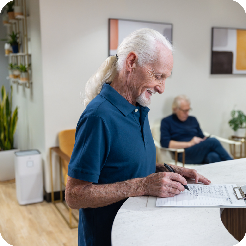 A patient smiling filling out paper work at a doctors office  filled with chairs, plants and a air purifier.