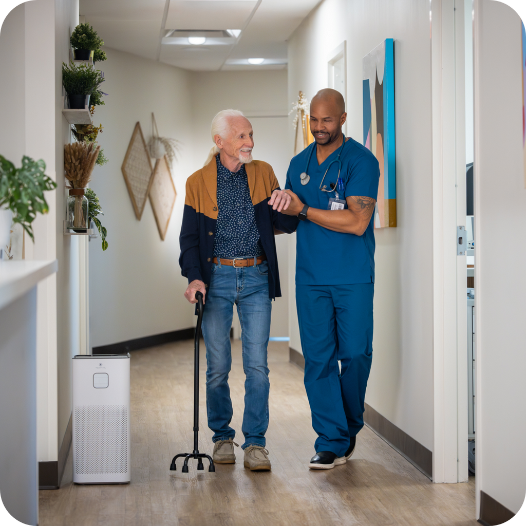a nurse practitioner helping a patient walk in the hallway of a hospital next to a air purifier and a reception desk. 